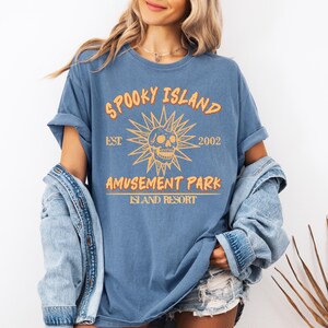 May include: Blue t-shirt with the text "SPOOKY ISLAND AMUSEMENT PARK ISLAND RESORT" in orange. The design includes a skull and sunburst graphic. The shirt is a relaxed fit.