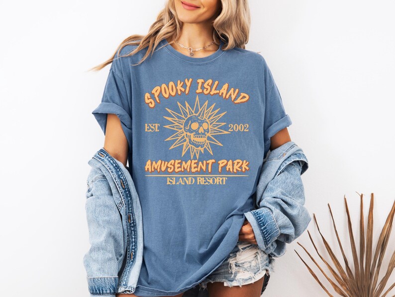 May include: Blue t-shirt with the text "SPOOKY ISLAND AMUSEMENT PARK ISLAND RESORT" in orange. The design includes a skull and sunburst graphic. The shirt is a relaxed fit.