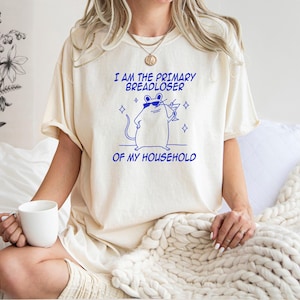 May include: An ivory t-shirt featuring a blue graphic of a mouse wearing sunglasses and holding a cocktail glass. The text above the mouse reads I AM THE PRIMARY BREADLOSER and the text below the mouse reads OF MY HOUSEHOLD in blue lettering.