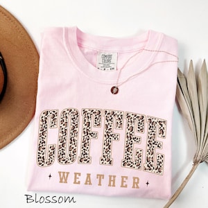 May include: A pale pink Comfort Colors t-shirt featuring the words "COFFEE WEATHER" in a leopard print design. A gold necklace with a pendant is on the shirt. A brown hat is on the left side of the image.