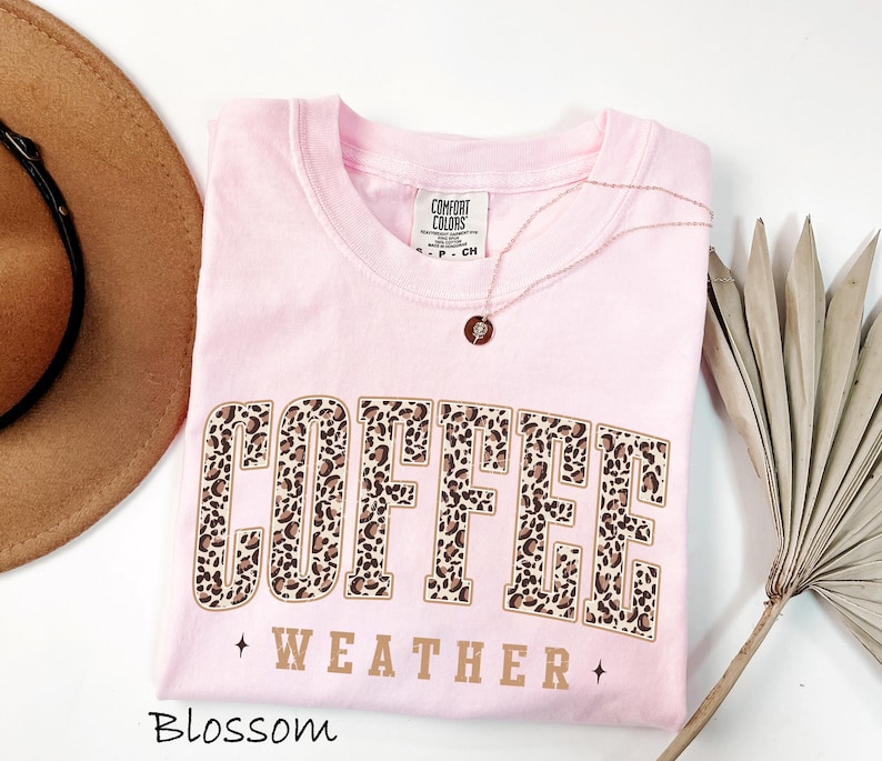 May include: A pale pink Comfort Colors t-shirt featuring the words "COFFEE WEATHER" in a leopard print design. A gold necklace with a pendant is on the shirt. A brown hat is on the left side of the image.