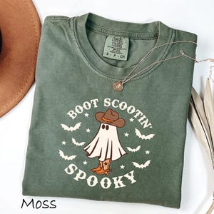 May include: A moss green t-shirt with a graphic of a ghost wearing a cowboy hat and boots, surrounded by bats and stars. The text reads "Boot Scootin' Spooky". A silver necklace with a pendant is draped on the shirt.