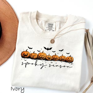 May include: Ivory-colored t-shirt featuring a row of carved orange pumpkins with black bat silhouettes above. The phrase "spooky season" is written in black script below the pumpkins. A gold necklace is draped on the shirt.