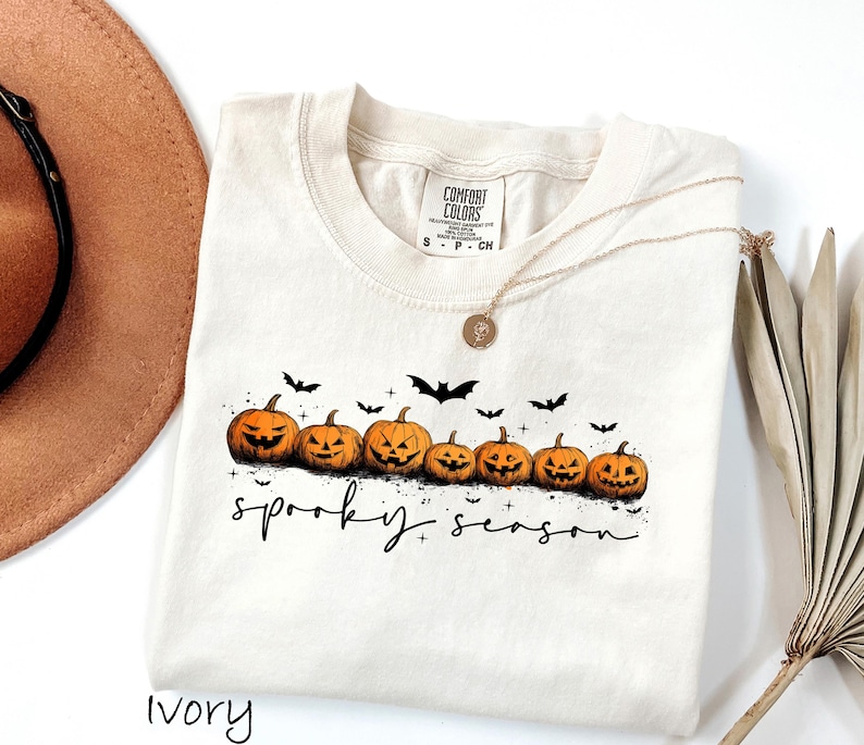 May include: Ivory-colored t-shirt featuring a row of carved orange pumpkins with black bat silhouettes above. The phrase "spooky season" is written in black script below the pumpkins. A gold necklace is draped on the shirt.