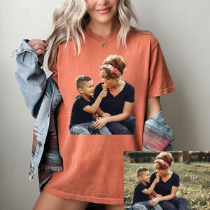 May include: Orange-red t-shirt featuring a printed image of a mother and child sharing an ice cream cone. The child is offering the ice cream to the mother. The shirt is a comfortable, casual style, suitable for everyday wear.