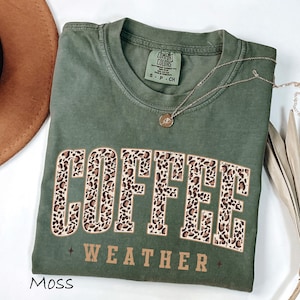May include: A moss green t-shirt with the words "COFFEE WEATHER" in a leopard print design. The shirt has a crew neck and short sleeves. A gold necklace with a pendant is draped on the shirt.