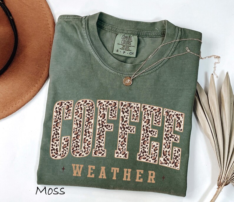 May include: A moss green t-shirt with the words "COFFEE WEATHER" in a leopard print design. The shirt has a crew neck and short sleeves. A gold necklace with a pendant is draped on the shirt.