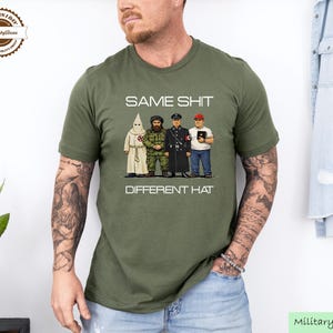 May include: Military green t-shirt featuring the text "SAME SHIT DIFFERENT HAT" above a graphic of four figures in different uniforms. The t-shirt has a crew neck.