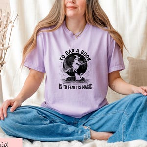 A woman wearing a purple t-shirt with a graphic design sits cross-legged on a bed, smiling.