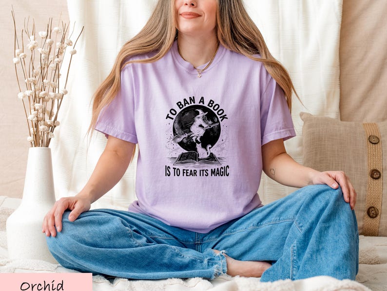 A woman wearing a purple t-shirt with a graphic design sits cross-legged on a bed, smiling.
