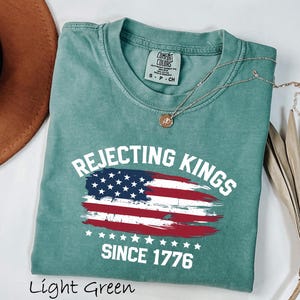 May include: Light green t-shirt with the text "Rejecting Kings Since 1776" and a distressed American flag design. The shirt is made of a soft material and has a classic crew neck.