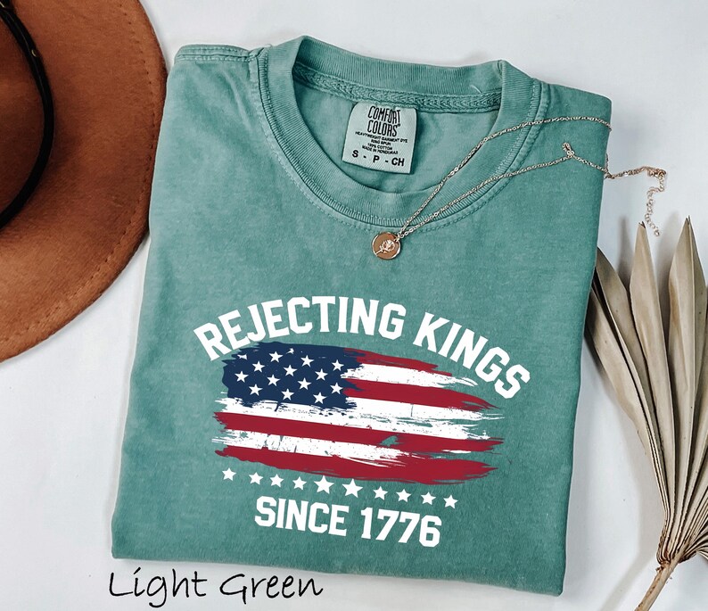May include: Light green t-shirt with the text "Rejecting Kings Since 1776" and a distressed American flag design. The shirt is made of a soft material and has a classic crew neck.