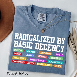 May include: Blue jean colored t-shirt with the text "RADICALIZED BY BASIC DECENCY" in white block letters. Below the text are colorful bars with words like "EMPATHY", "JUSTICE", and "LGBTQ+ RIGHTS". A gold necklace is draped over the shirt.