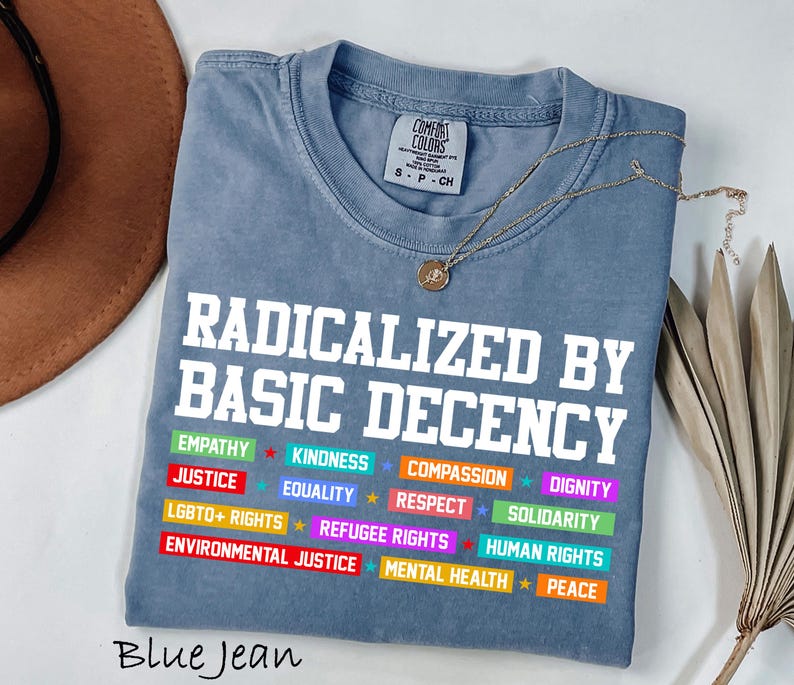 May include: Blue jean colored t-shirt with the text "RADICALIZED BY BASIC DECENCY" in white block letters. Below the text are colorful bars with words like "EMPATHY", "JUSTICE", and "LGBTQ+ RIGHTS". A gold necklace is draped over the shirt.