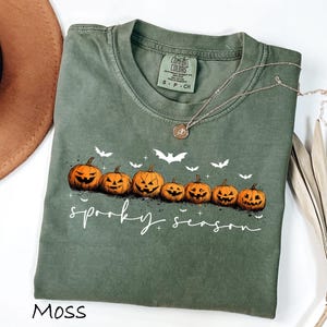 May include: Moss green t-shirt with a crew neck featuring a row of jack-o'-lanterns and bats. The words "spooky season" are written in white script. A gold necklace is draped across the shirt.