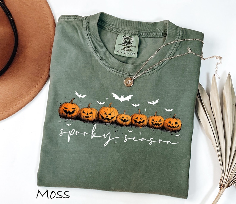 May include: Moss green t-shirt with a crew neck featuring a row of jack-o'-lanterns and bats. The words "spooky season" are written in white script. A gold necklace is draped across the shirt.