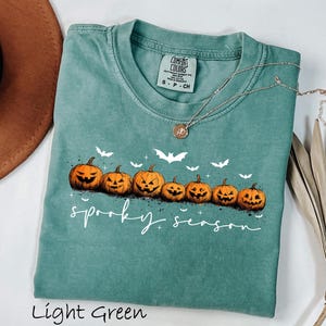 May include: Light green t-shirt with a crew neck featuring a row of carved orange pumpkins and white bats. The words "spooky season" are written in a script font. A silver necklace with a pendant is draped on the shirt.