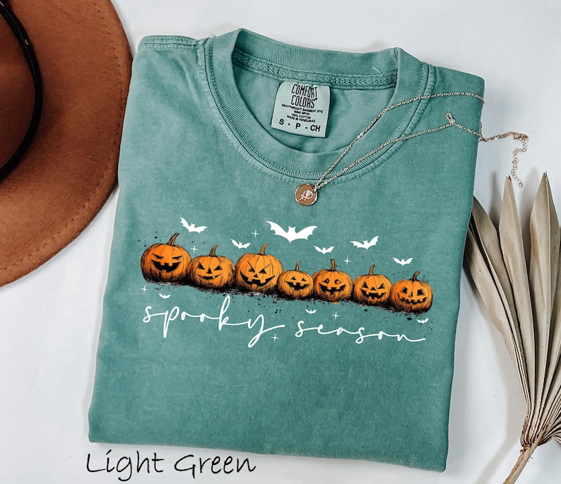 May include: Light green t-shirt with a crew neck featuring a row of carved orange pumpkins and white bats. The words "spooky season" are written in a script font. A silver necklace with a pendant is draped on the shirt.