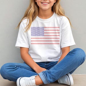 A young woman wearing a white t-shirt with an American flag design is sitting cross-legged on the ground, smiling at the camera.