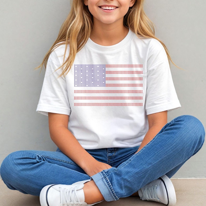 A young woman wearing a white t-shirt with an American flag design is sitting cross-legged on the ground, smiling at the camera.
