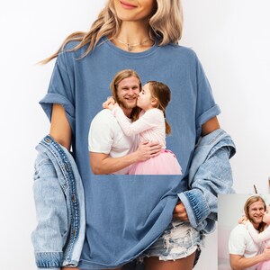 May include: A blue t-shirt featuring a photograph of a man and a young girl embracing and kissing. The person wearing the shirt is also wearing a denim jacket and jean shorts. The shirt is a comfortable, casual style.