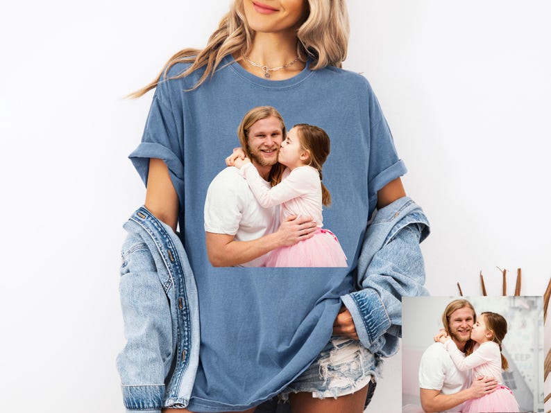 May include: A blue t-shirt featuring a photograph of a man and a young girl embracing and kissing. The person wearing the shirt is also wearing a denim jacket and jean shorts. The shirt is a comfortable, casual style.
