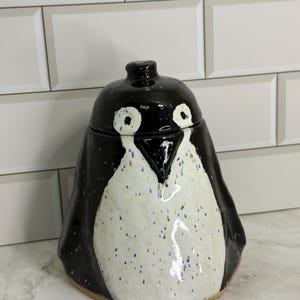 May include: A ceramic jar shaped like a penguin with a removable lid. The penguin has a glossy black exterior, a white chest area with small colorful speckles, and two white circular eye patches surrounding black painted eyes.