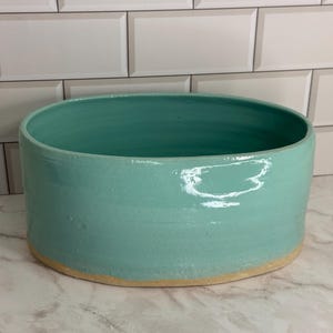 May include: A wide, cylindrical ceramic bowl with a smooth, glossy turquoise glaze on the interior and exterior. The base of the bowl is unglazed, revealing a natural, light tan clay texture that contrasts with the vibrant blue-green finish.