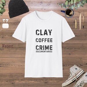Op de afbeelding: Wit t-shirt met de woorden "CLAY COFFEE CRIME DOCUMENTARIES" in zwarte, verweerde letters. Het shirt wordt gepresenteerd op een houten oppervlak, met een zwarte muts, oortelefoons en een paar hoge sneakers.