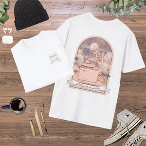Peut inclure: T-shirt blanc avec un graphique d'un squelette travaillant sur un tour de potier. Le graphique est de style vintage avec une palette de couleurs brun et beige. Le texte sur le graphique dit "I'll Death do us Part!"