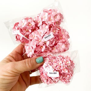 May include: Two packages of pink paper flowers with white centers. The flowers are in clear plastic bags. Each bag has a white label with the text "SAA-579" and "SAA-564".