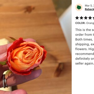 May include: A close-up of a single orange and red artificial rose bud. The rose is held in a hand with a silver ring. The background is a wooden surface.