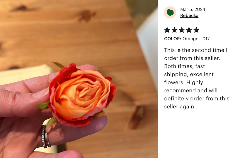 May include: A close-up of a single orange and red artificial rose bud. The rose is held in a hand with a silver ring. The background is a wooden surface.