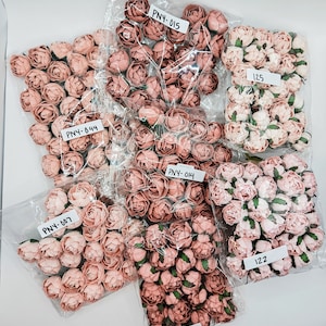 May include: Multiple clear plastic bags containing artificial flowers in shades of pink and peach. Each bag has a white label with a product code. The flowers resemble small roses, suitable for crafts or decoration. The bags are arranged on a white surface.