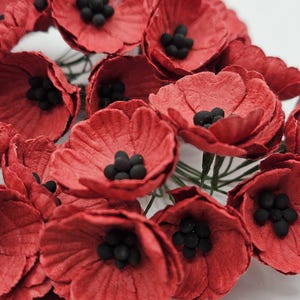 May include: Close-up of a cluster of red paper poppy flowers with black centers. The flowers have textured petals and green stems, creating a vibrant and detailed floral arrangement. Ideal for crafting or decorative purposes.