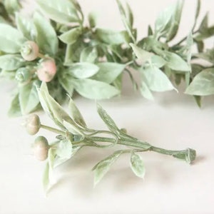 May include: Close-up of artificial greenery with light green leaves and small, round, pinkish-tan berries. The stems are a muted green color. The leaves have a soft, textured appearance. The background is a light, neutral color.