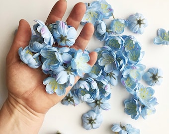 May include: A handful of light blue artificial flowers with a yellow centre. The flowers are scattered on a white surface.