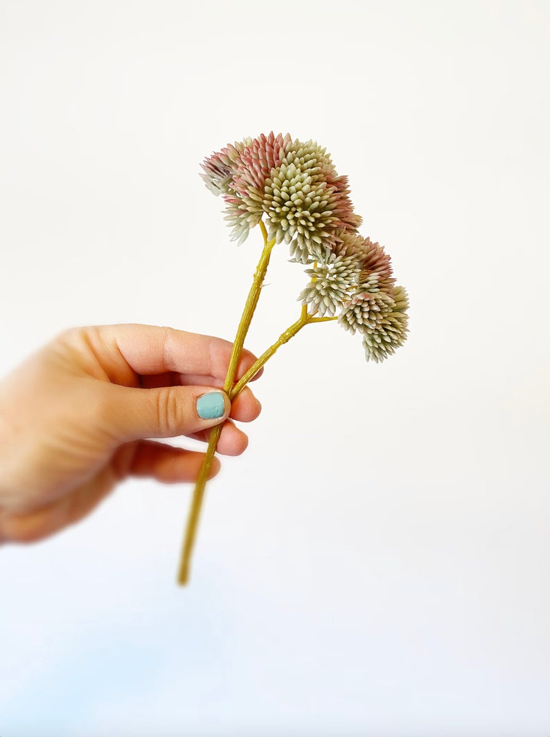 以下が含まれることがあります： 緑とピンクのふわふわした花頭を持つ、人工の花の茎が3本束ねられたブーケ。茎は薄い茶色です。