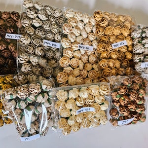 Op de afbeelding: Assortiment van zes zakjes met kleine papieren bloemen in verschillende kleuren, waaronder bruin, beige, geel en crème. De zakjes zijn gelabeld met de productcode en de hoeveelheid.