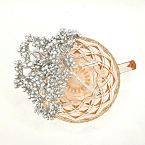 Puede incluir: Una taza de t&eacute; de cristal rosa p&aacute;lido con un patr&oacute;n geom&eacute;trico de diamantes en el exterior y un asa lateral. Dentro de la taza hay un racimo de peque&ntilde;as bayas artificiales de color plata met&aacute;lico unidas a tallos de alambre fino y flexible.