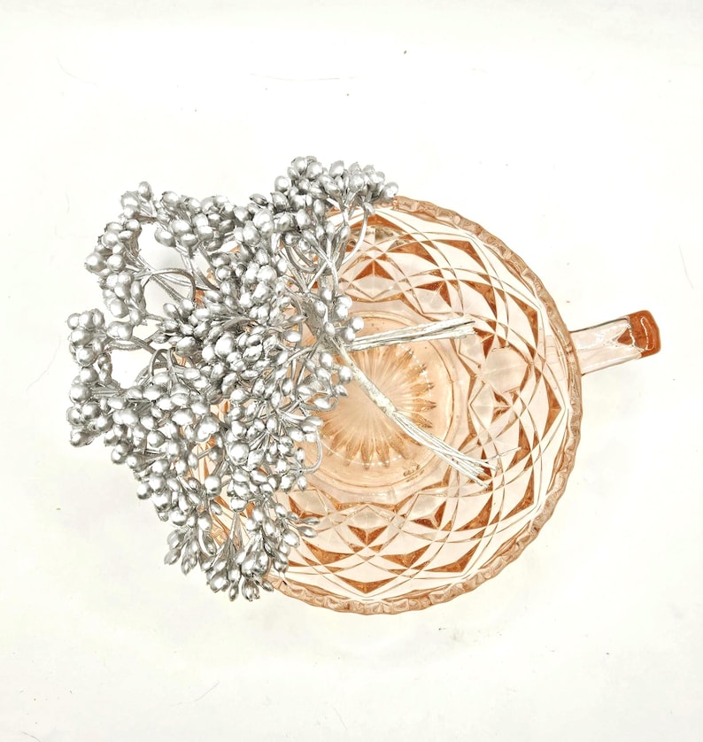 Puede incluir: Una taza de t&eacute; de cristal rosa p&aacute;lido con un patr&oacute;n geom&eacute;trico de diamantes en el exterior y un asa lateral. Dentro de la taza hay un racimo de peque&ntilde;as bayas artificiales de color plata met&aacute;lico unidas a tallos de alambre fino y flexible.