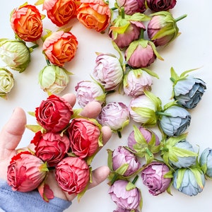 May include: Assortment of artificial flower buds in various colors, including peach, pink, red, and blue. The buds are arranged on a white surface.