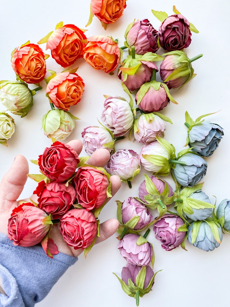 May include: Assortment of artificial flower buds in various colors, including peach, pink, red, and blue. The buds are arranged on a white surface.