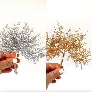 May include: Two bunches of artificial greenery, one silver and one gold, with thin stems. The greenery is arranged in a bushy, tree-like shape.