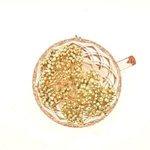 Puede incluir: Una taza de t&eacute; de cristal rosa p&aacute;lido con un patr&oacute;n de diamantes facetados en el exterior, rellena con un racimo de peque&ntilde;as bayas decorativas de color dorado met&aacute;lico sobre tallos finos. La taza tiene un asa en el lado derecho.