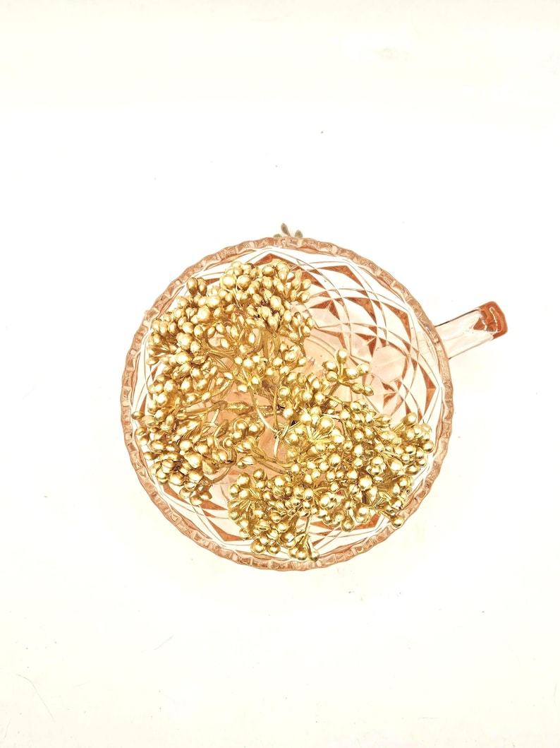 Puede incluir: Una taza de t&eacute; de cristal rosa p&aacute;lido con un patr&oacute;n de diamantes facetados en el exterior, rellena con un racimo de peque&ntilde;as bayas decorativas de color dorado met&aacute;lico sobre tallos finos. La taza tiene un asa en el lado derecho.