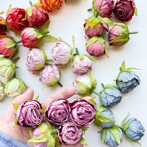 May include: Assortment of artificial flower buds in various colors, including pink, purple, blue, orange, and red. The buds are arranged on a white surface and some are held in a hand.