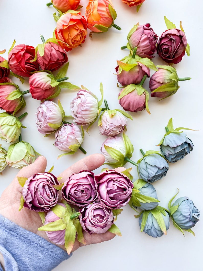May include: Assortment of artificial flower buds in various colors, including pink, purple, blue, orange, and red. The buds are arranged on a white surface and some are held in a hand.