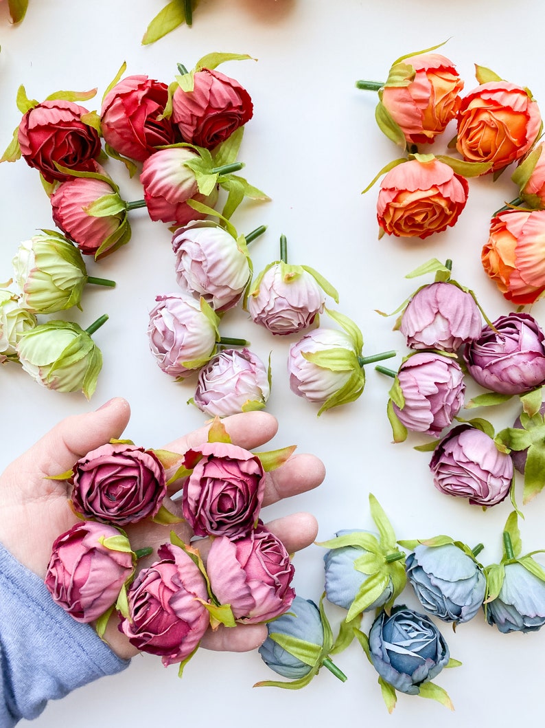 May include: Assortment of artificial flower buds in various colors, including pink, purple, blue, orange, and red. The flower buds are arranged on a white surface.