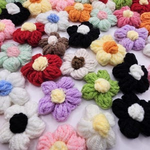 May include: A collection of handmade yarn flowers in various colors, including pink, white, black, yellow, and purple. Each flower has a contrasting center, creating a textured and colorful display. These floral embellishments are ideal for crafts and decorations.